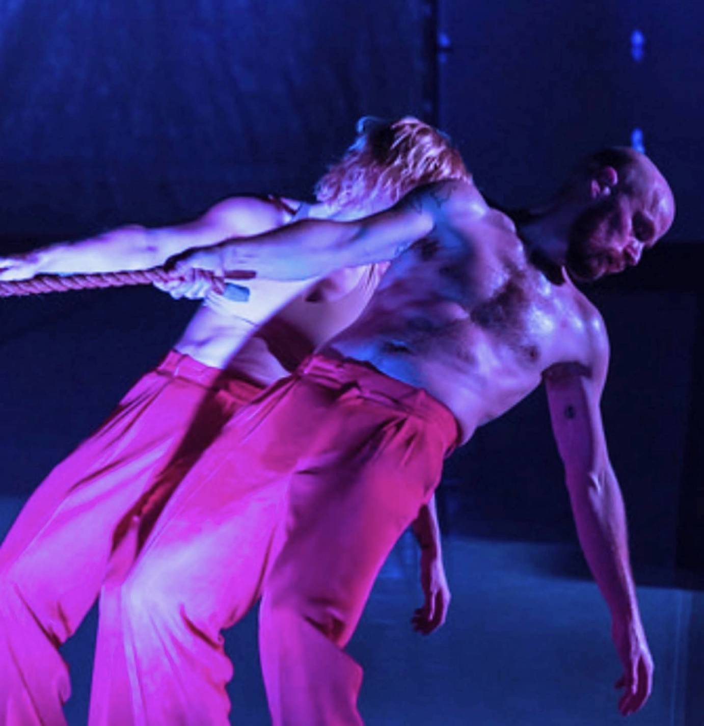 A male performer, chest bare, wearing long pants is compressed with his partner, her body behind his. Both are wearing bright pink long pants. They are leaning to their right grasping a rope. The color of the photo is purplish.